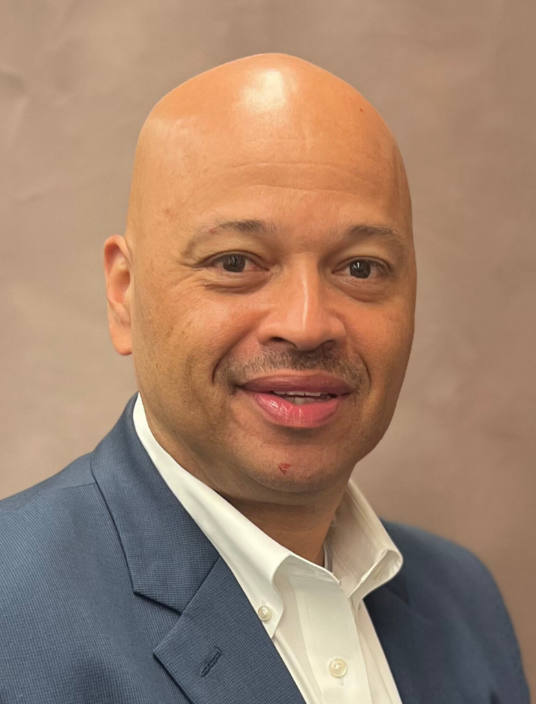 A bald man with a faint mustache and goatee wearing a navy blue blazer and white shirt smiles while looking at the camera. The background is a neutral brown color.